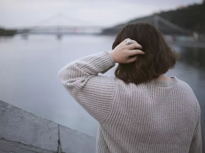 Žena stojí u řeky s výhledem na městský most v oblačném počasí, zadní pohled. Osamělá žena za zamračeného dne chodí po městě.